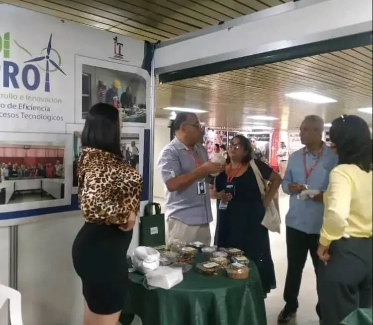 Fotografía interior del un stand del CEPROT de la Universidad de Las Tunas en el ExpoCaribe en Cuba. Un stand exhibe materiales promocionales, incluyendo una pancarta que dice "CEPROT" (Un proyecto de desarrollo e Innovación en energías renovables de la Universidad de Las Tunas) junto con texto en español que hace referencia a la innovación y la eficiencia en los procesos tecnológicos. Varias personas están reunidas en una mesa llena de diversos recipientes que parecen ser pequeños alimentos o refrigerios deshidratados. Una mujer con cabello largo y oscuro, una blusa con estampado de leopardo y una falda negra está de pie junto a la mesa. Varios hombres y mujeres conversan, especialmente alrededor de la mesa con refrigerios. Los hombres visten camisas abotonadas y las mujeres, diversas blusas y faldas. El fondo sugiere una sala de convenciones al aire libre o un espacio similar con una estructura de techo visible. Las paredes del stand son blancas.