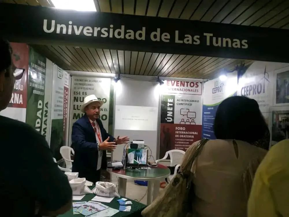 Un hombre con traje oscuro y sombrero claro da una presentación en un stand del CEPROT de la Universidad de Las Tunas. El stand está decorado con carteles y pancartas. Algunos carteles tienen texto en español, incluyendo el nombre de la universidad: "Universidad de Las Tunas". Otros carteles mencionan "Eventos Internacionales", "Únete" y "Foro Internacional de Oratoria". Varias personas entre el público escuchan la presentación. Frente al presentador hay una mesa con papeles y pequeños objetos. La sala tiene un techo beige claro o tostado. La impresión general es la de un evento académico profesional y organizado.